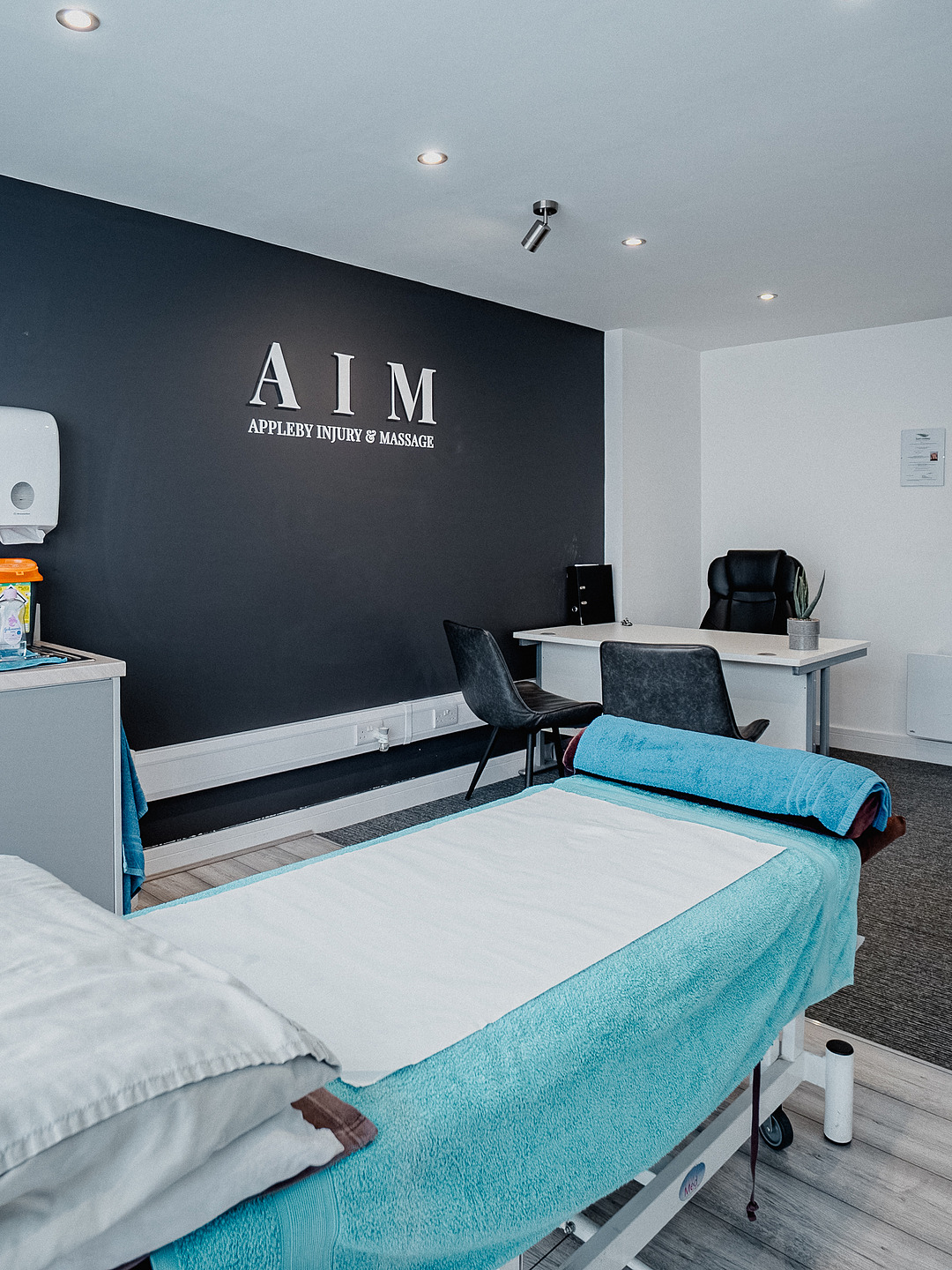 The treatment room at Appleby Injury & Massage, with a made-up couch below the AIM signage and the consultation desk beyond.