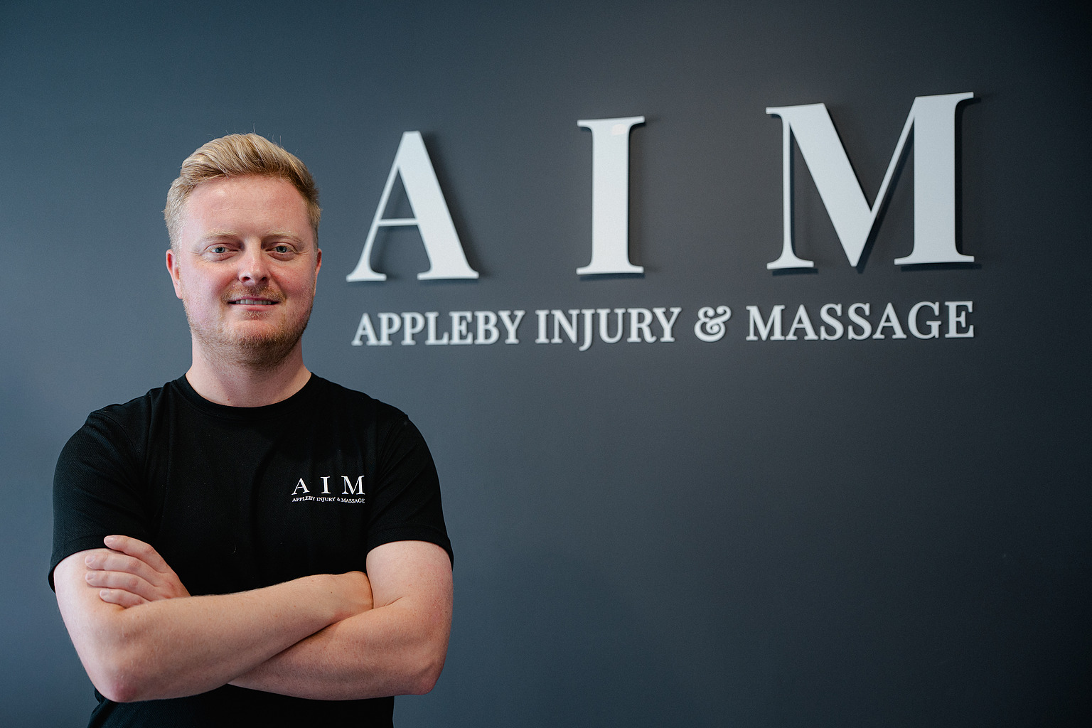 William Appleby, BSc Hons Sports Therapist, in front of the AIM signage at the clinic.